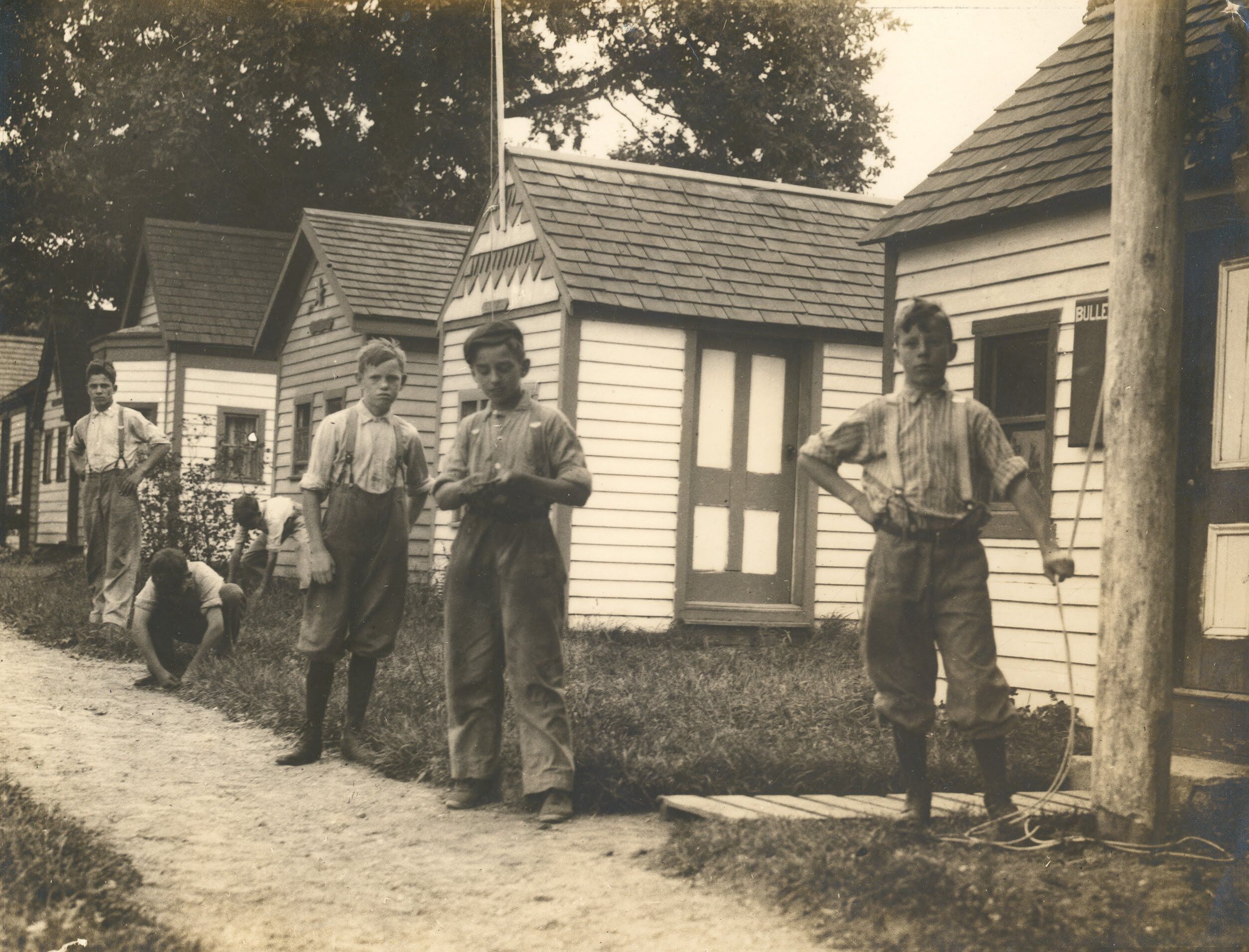 The image shows a group of young boys standing in front of a row of small, simple houses. The boys are dressed in work clothes, and some are holding tools or ropes. The houses appear to be basic structures, possibly for workers or a camp. The scene has a vintage feel.